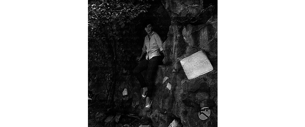 Barbara Zacconi seduta in una piccola grotta scavata nella roccia; nella foto compare un pannello di marmo con la scritta 'Casa di Pietro Benvenuti' - totale