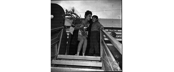 Vera Tschechowa ripresa in cima alle scalette dell'aereo con fiori in mano ed un bambino accanto - totale