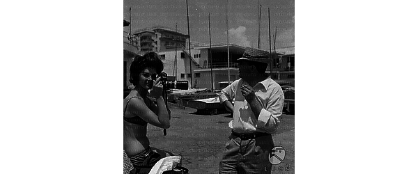 Francine Royer in bikini in un molo punta l'obiettivo di una macchina fotografica; accanto a lei un uomo - piano americano