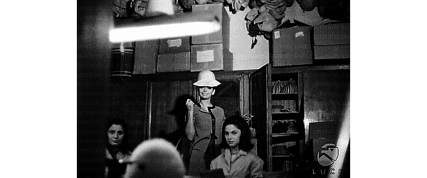 Barbara Steele prova un modello in un negozio di cappelli. Due lavoranti sedute al tavolo davanti a lei - campo medio