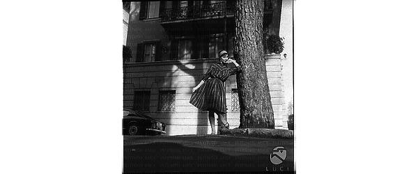 Una modella con un vestito elegante in posa in una aiuola vicino ad un albero. Campo medio