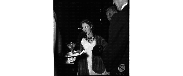 Un'attrice con un a stola di pelliccia  che prende un bicchiere di aperitivo. Piano americano
