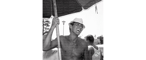 Walter Chiari a torso nudo sul set del film: sorridente, con cappellino bianco e un ombrellone parasole in mano
