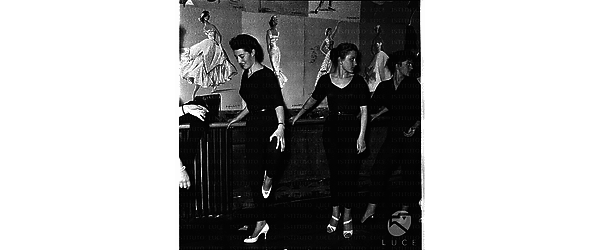 Tre ragazze colte alla sbarra durante una lezione di danza. Cartoni raffiguranti modelli d'alta moda sono affissi alla parete di fondo della sala di danza. Totale