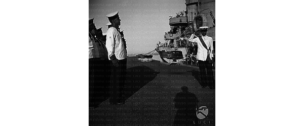 Un alto ufficiale passa in rassegna un gruppo di marinai schierati sul molo di un porto, lato si vede la nave Bersagliere attraccata