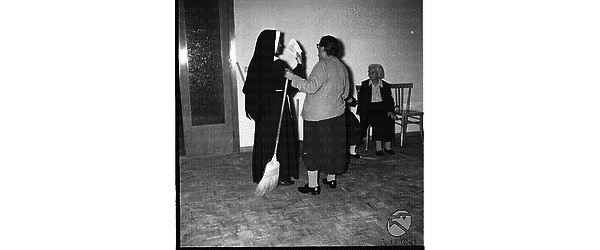 Una suora mostra dei fogli di carta ad una donna anziana con la scopa; campo medio