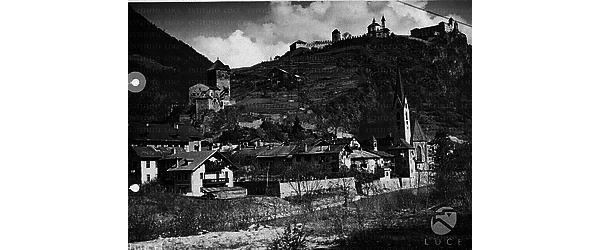 Veduta di un paese (torre medievale, case, chiesa con campanile) che si estende lungo il corso di un torrente in una vallata; in cima ad un monte si vede un muro merlato ed una chiesa