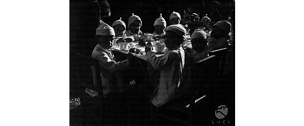 I bambini del Dispensario in posa a tavola durante la colazione. Essi indossano cappellino e camice bianco - Totale