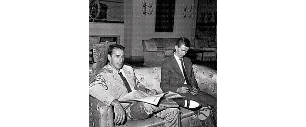Roma Bettini seduto su un divano della hall di un albergo, con un quotidiano; al suo fianco un uomo; interno